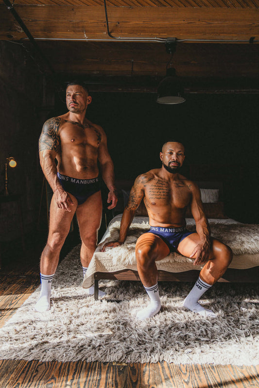 Two men in a bedroom wearing navy blue MANBUNS underwear—one in briefs standing, one in boxer briefs seated—both styled with white MB crew socks featuring black lettering and stripes.