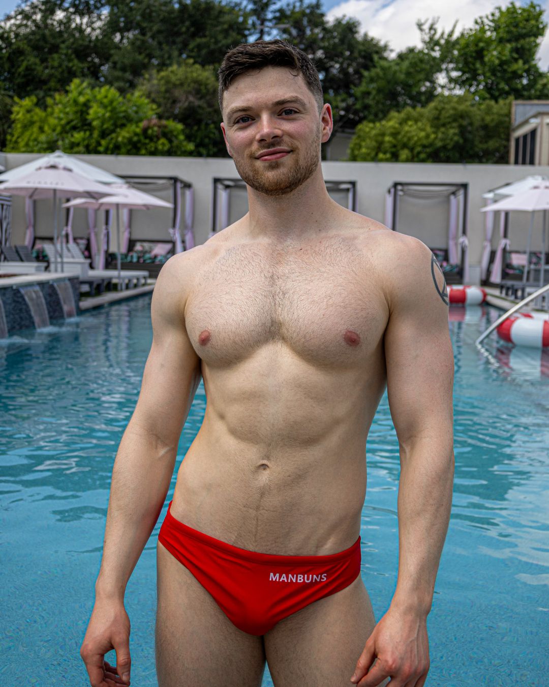man standing at the pool shirtless wearing a red swim brief with the brand logo on the right hip