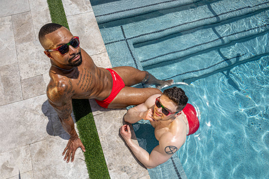 two men at the pool wearing rainbow sunglasses that say manbuns on the left side. photo is taken upward and both models are looking up
