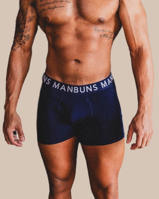 Man wearing navy blue boxer briefs with 'MANBUNS' waistband on a beige background