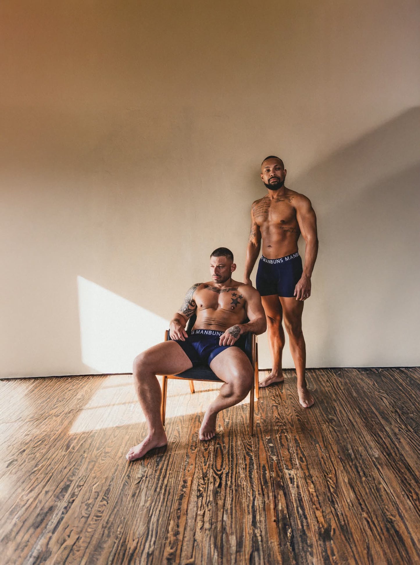 two models wearing midnight navy blue boxer briefs manbuns underwear. one model is sitting in a chair and to the right of him the other model is standing. both are in front of a white blank wall with wooden floors. 