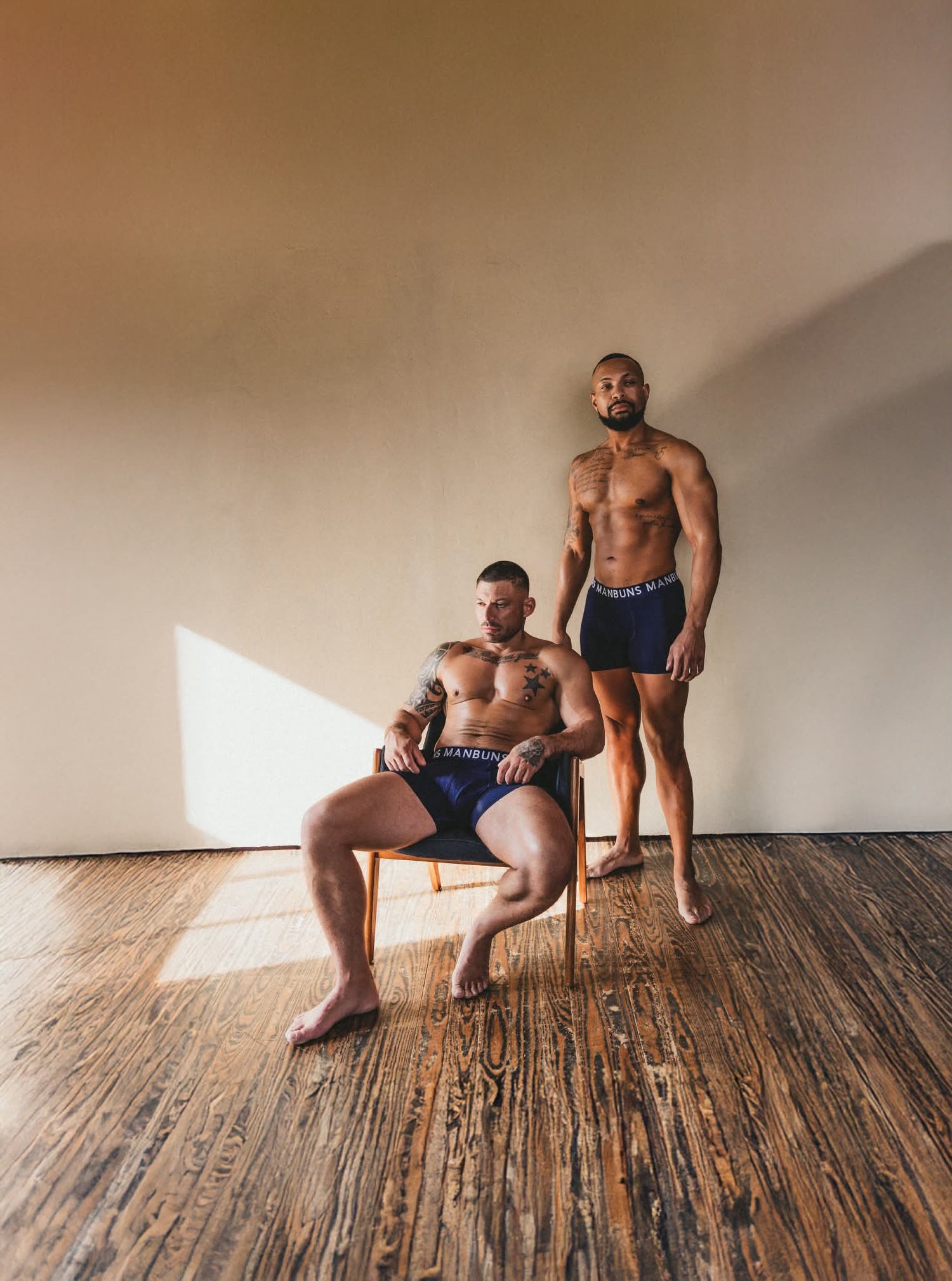 two models wearing midnight navy blue boxer briefs manbuns underwear. one model is sitting in a chair and to the right of him the other model is standing. both are in front of a white blank wall with wooden floors. 